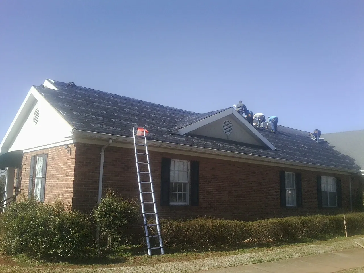 Crew tearing shingles off a brick ranch roof with a ladder set — Metal Roof Repair work by our crew in Casselton