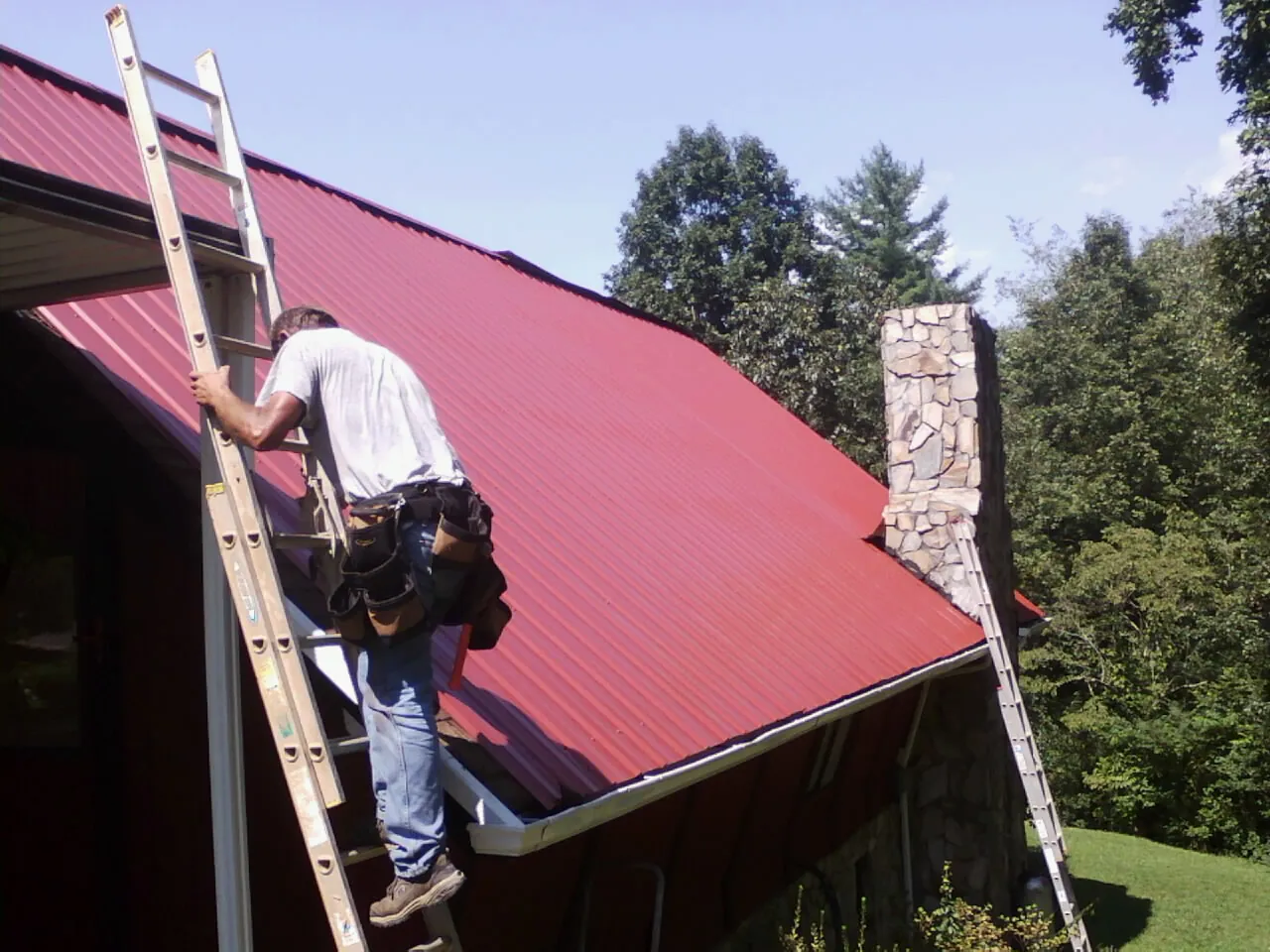 Roofer on a ladder beside a red metal roof and stone chimney — Soffit & Fascia Repair by Roofing Brothers in Casselton